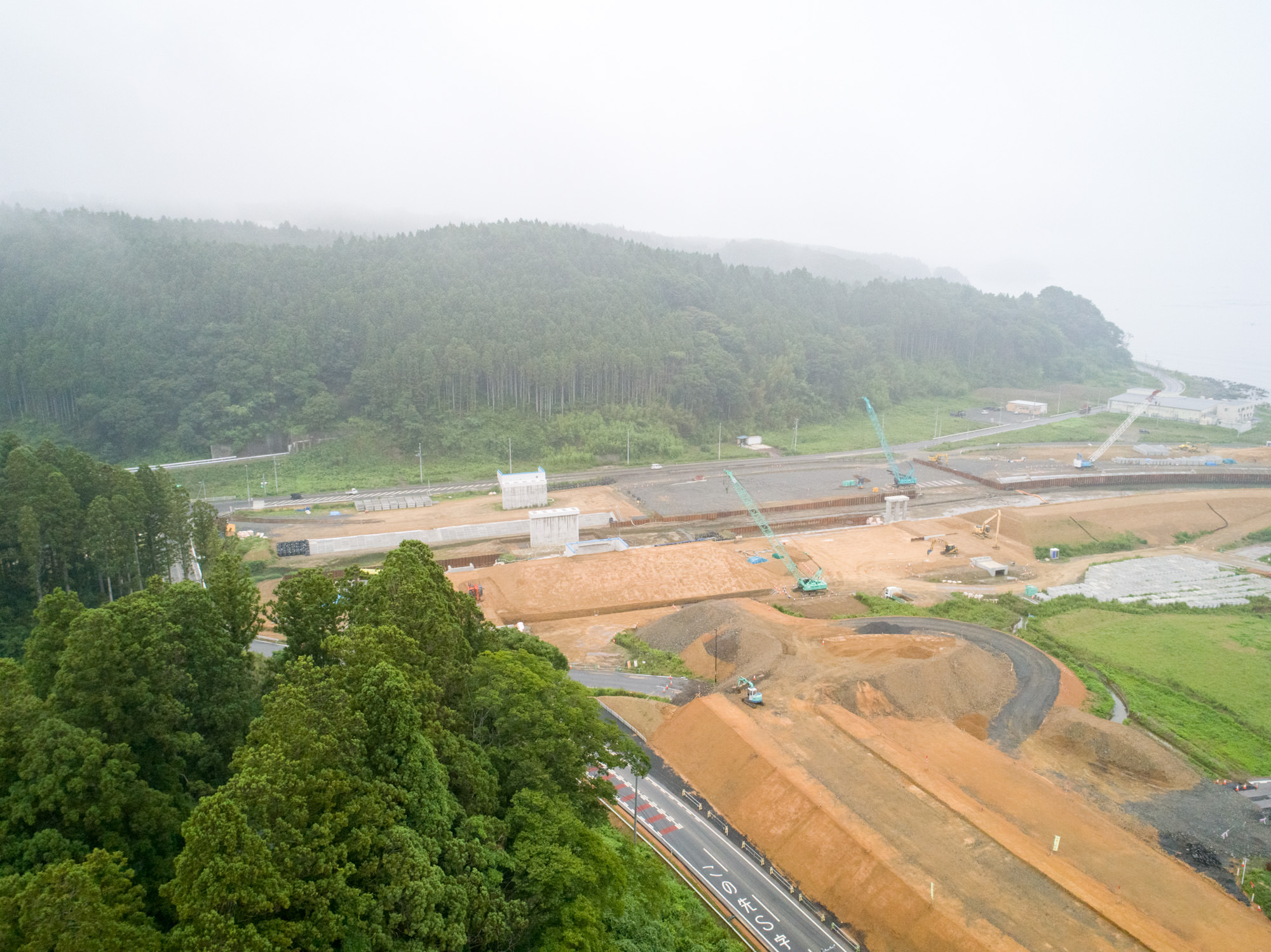 緑豊かな森林に囲まれた地域で、複数のクレーンや建設車両が稼働し、土の山や建設途中の構造物が点在する工事現場を捉えた、自然と開発が交差する風景の写真