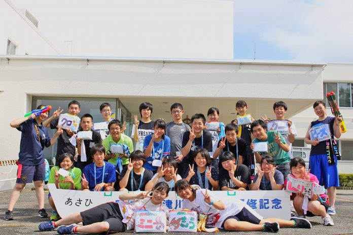 屋外で、手作りの名前札や水鉄砲を持った若者たち約25人が笑顔で集まり、ピースサインをしながら記念撮影をしている集合写真