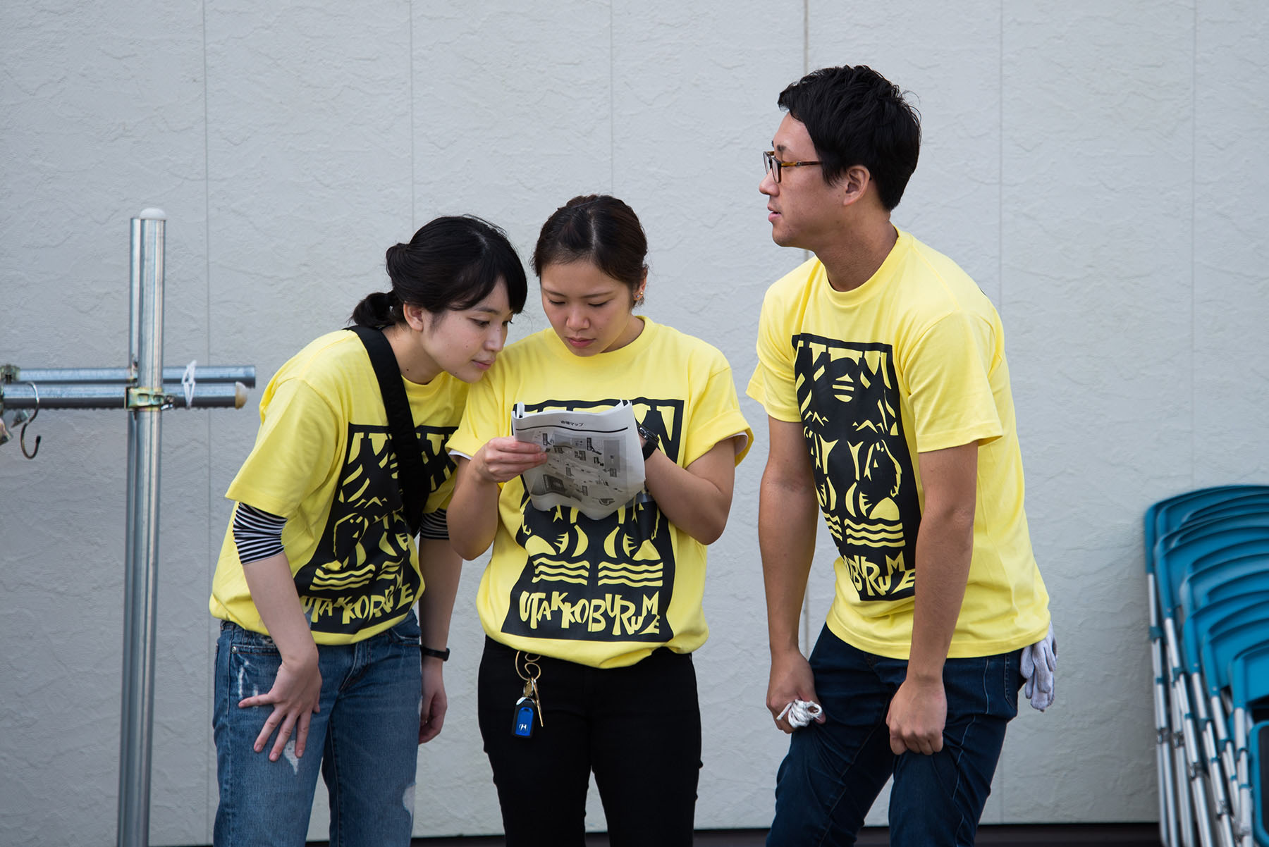 UTAKKOBURUMEの黄色いTシャツを着た若い男女3人が屋外で集まり、1人の女性が手に持つ紙を3人で真剣な表情で見ている写真