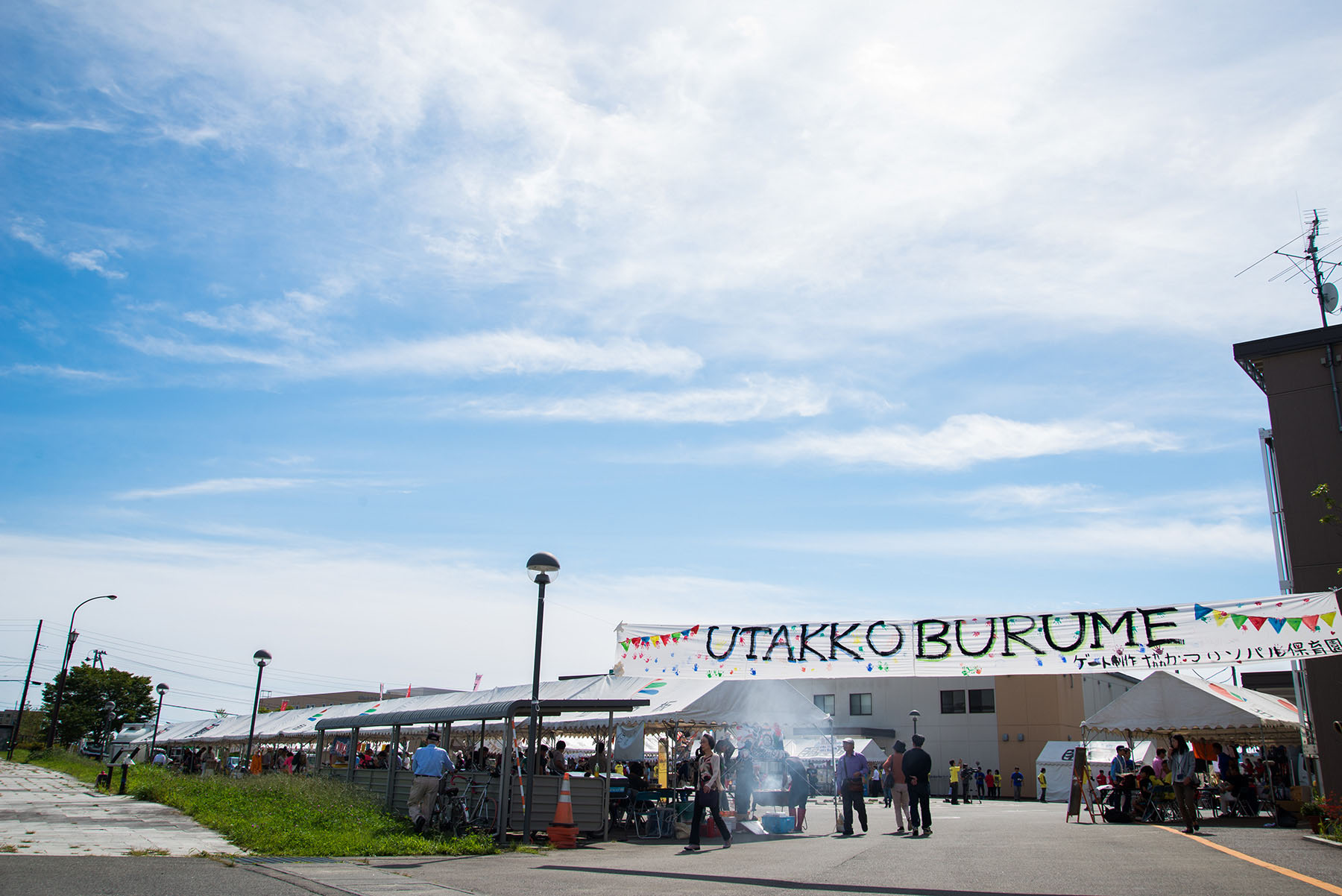 青空の下、「UTAKKOBURUME」と書かれた横断幕のある会場入り口にたくさんの人が集まり、テントの下でにぎわう様子が遠くから写されている写真