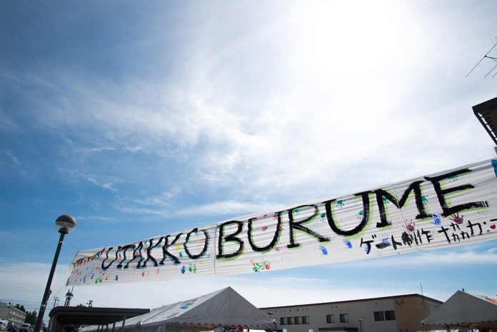 青空の下「UTAKKO BURUME」と手書きで書かれ、色とりどりの手形が押された横断幕が掲げられた、イベントゲートの写真