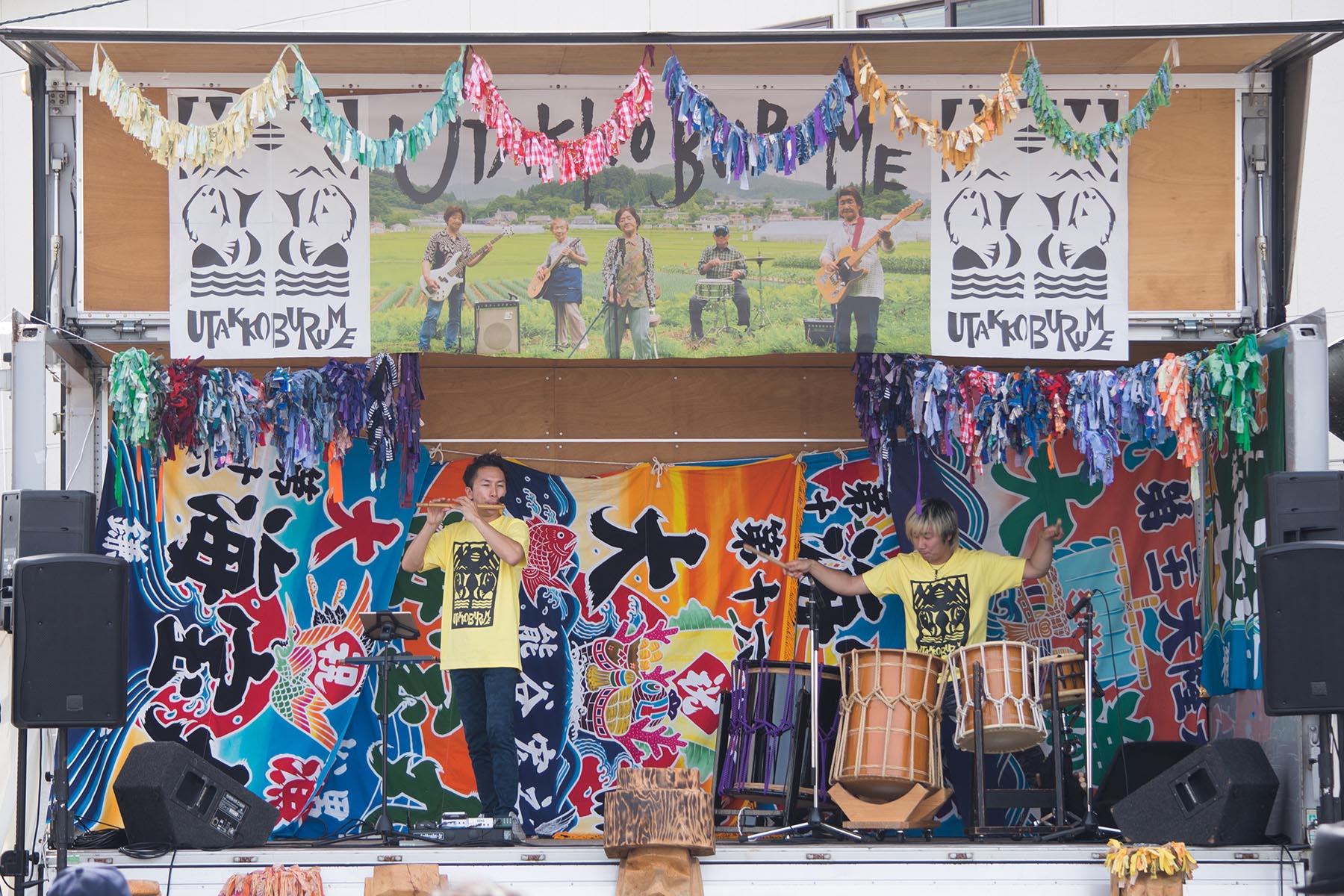 カラフルな大漁旗や布飾りで彩られた野外ステージ上で、黄色いTシャツを着た男性二人が横並びになり、一人は横笛を演奏しもう一人は和太鼓を叩いている写真