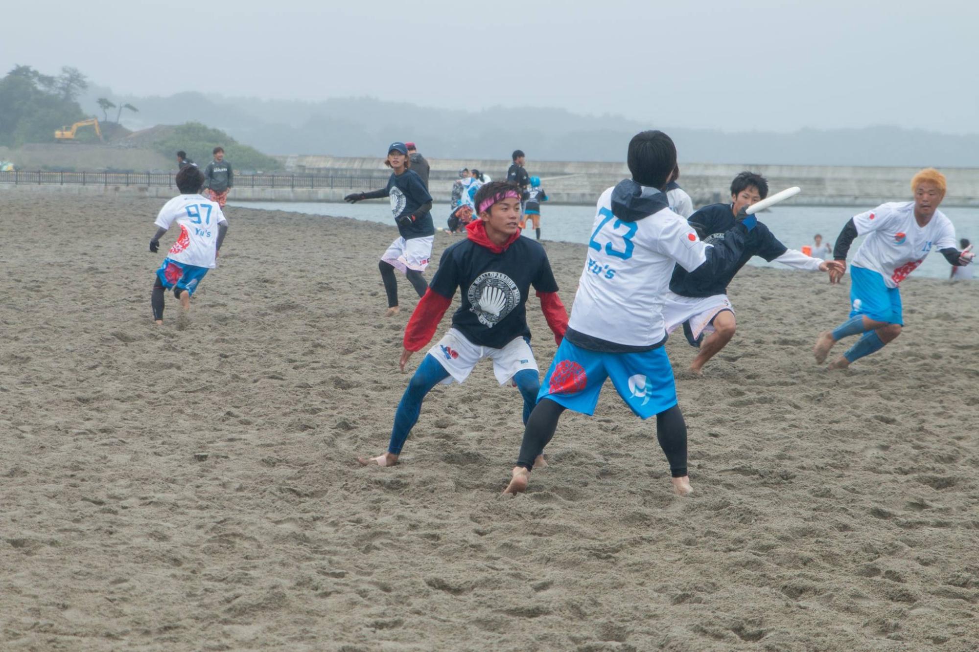 白い砂浜の上で選手たちが元気よく駆け回っている円盤型のフリスビーを使ったスポーツの写真