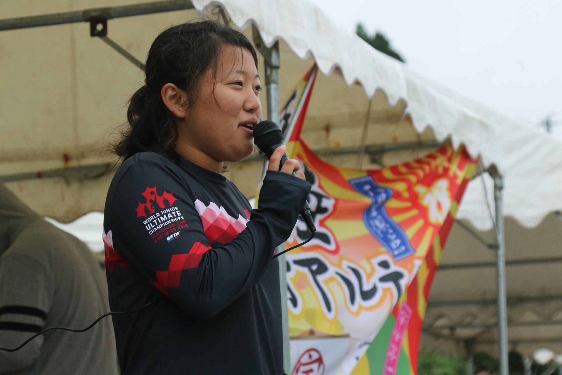 白いテントの前で女性の宮岡さんがマイクで挨拶する様子の写真