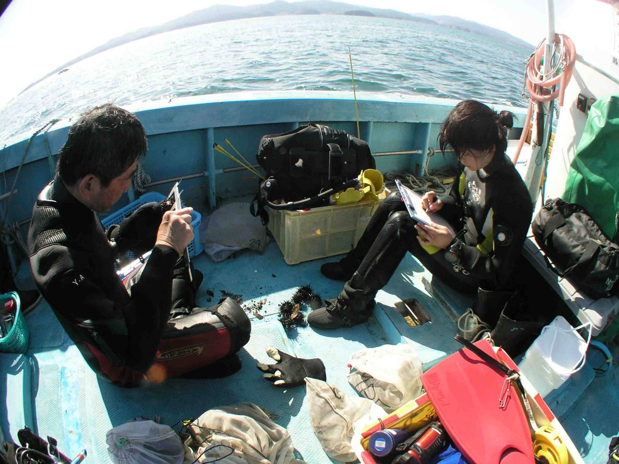 道具や容器が周囲に並ぶ青い漁船の上で、ウェットスーツを着た2人のダイバーが、採取したウニや海藻などのサンプルを手に観察・記録している海上調査の写真