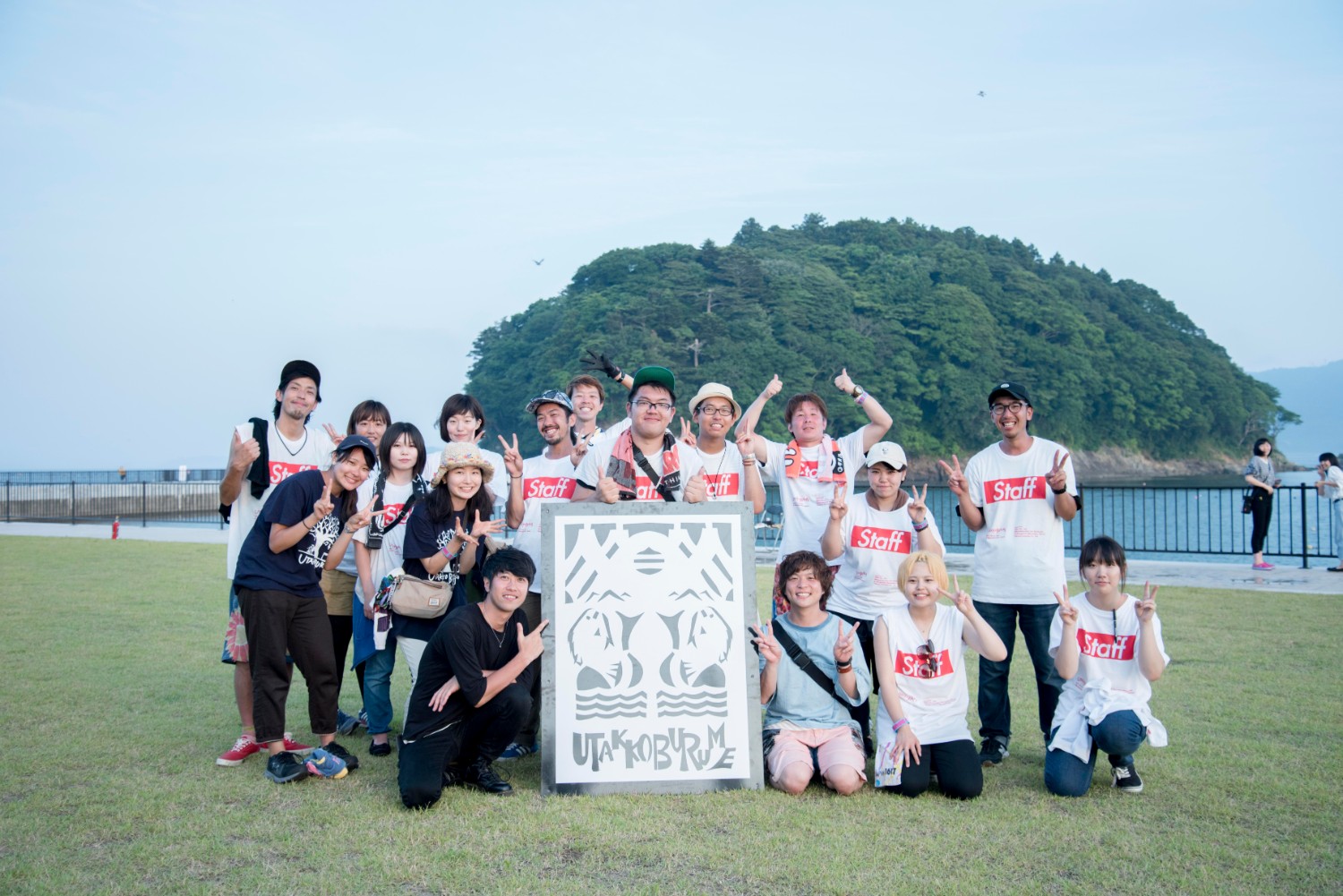 海辺の芝生の上で、笑顔の若者たちが「Staff」と書かれたTシャツを着て集合写真を撮っている写真