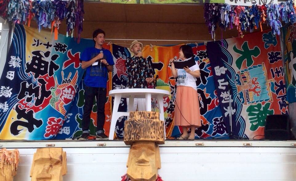 たくさんの大漁旗で飾られたステージの上に白い丸テーブルが置かれ、その後ろにマイクを持った2人の男性と女性リポーターが立って話をしている写真