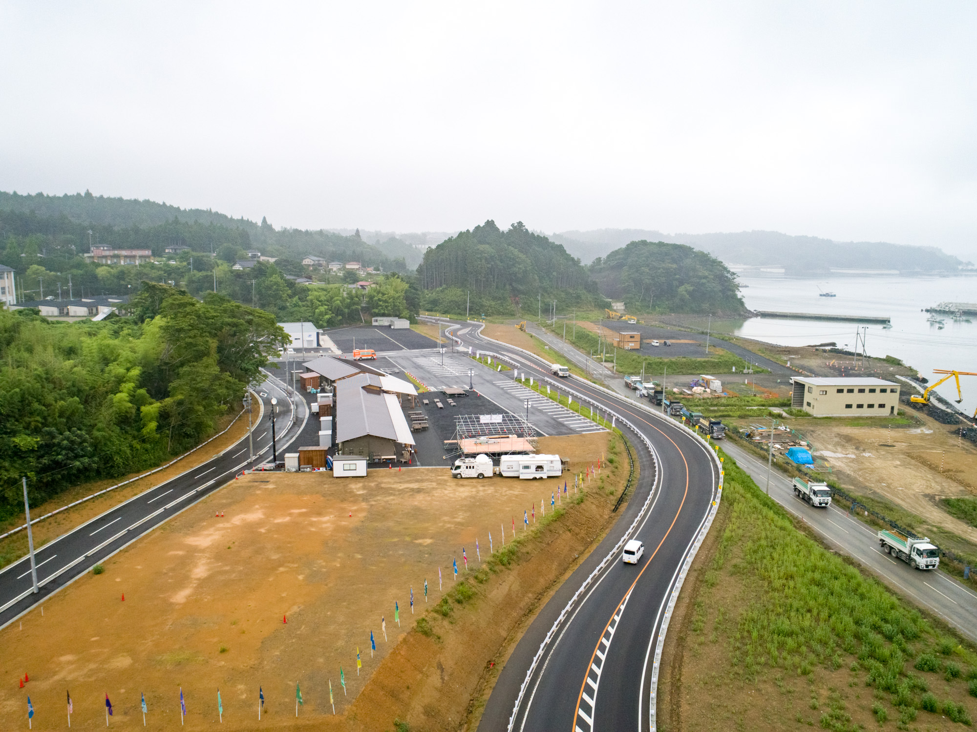 緑に囲まれた丘陵地帯で、傾斜屋根の大きな建物や複数の小さな構造物が建設中の工事現場を上空から捉え、舗装された道路には車両が走行・駐車しており、背景には水辺も広がる地域の開発風景の写真