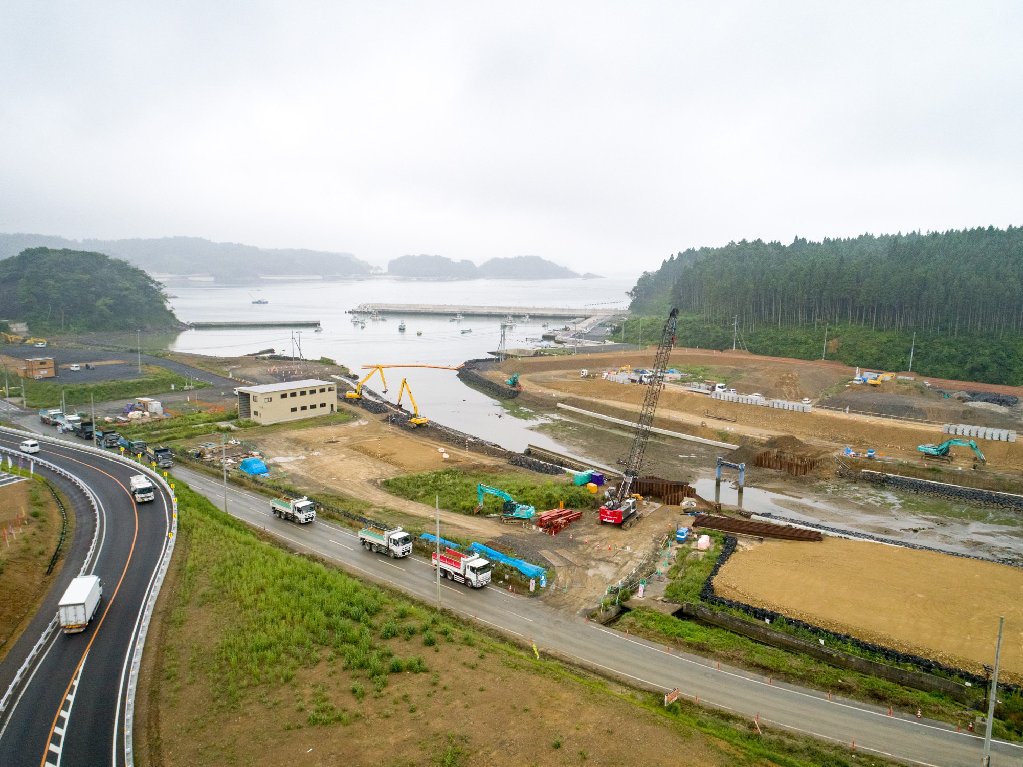 曇り空の下、港に隣接する工事現場で、クレーンやトラックなどの建設機材が稼働し、周囲の道路には車両が行き交い、背景には木々に覆われた丘と水上の船が見える、海辺の開発が進む様子を上空から捉えた写真