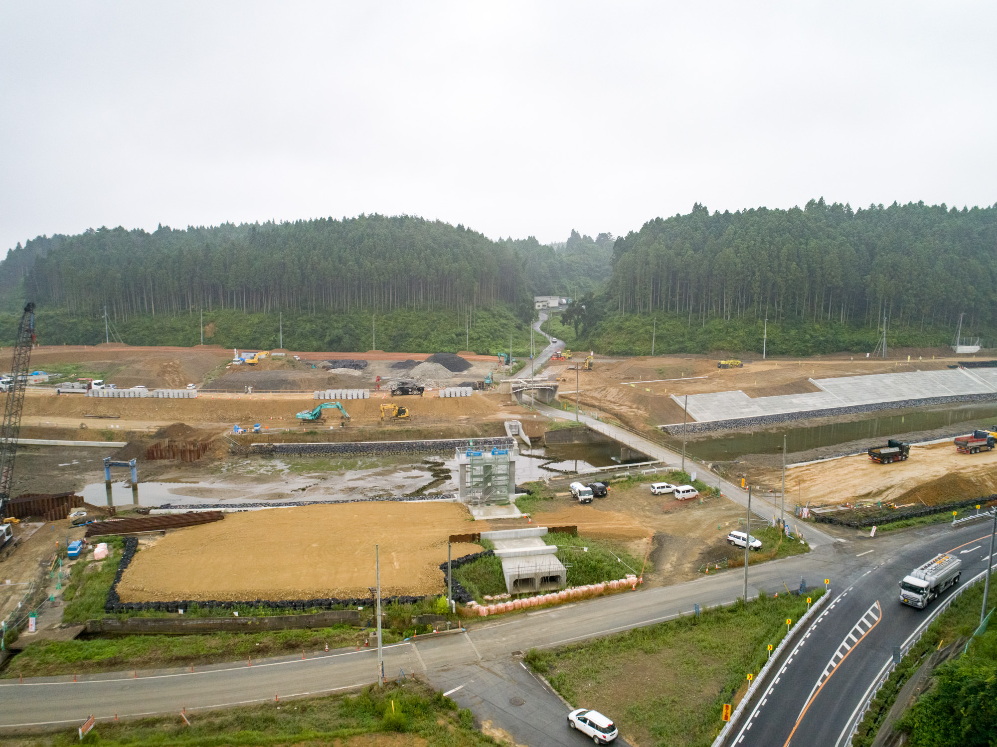 手前に道路が走り、その奥にはクレーンやショベルカー、トラックなどの建設機材が並ぶ整地された土地が広がり、いくつかの構造物が建設途中の様子を見せる、背景に森林が広がる開発初期の工事現場を上空から捉えた写真