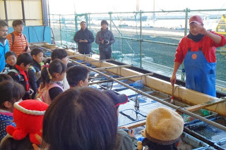 屋根のある水槽の前で赤い作業着の漁師が説明をし、子どもたちが熱心に話を聞いている様子の写真