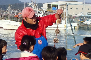 赤い作業着の漁師が子どもの貝が結び付けてあるロープを見学に来た子どもたちにみせながら説明をしている様子の写真