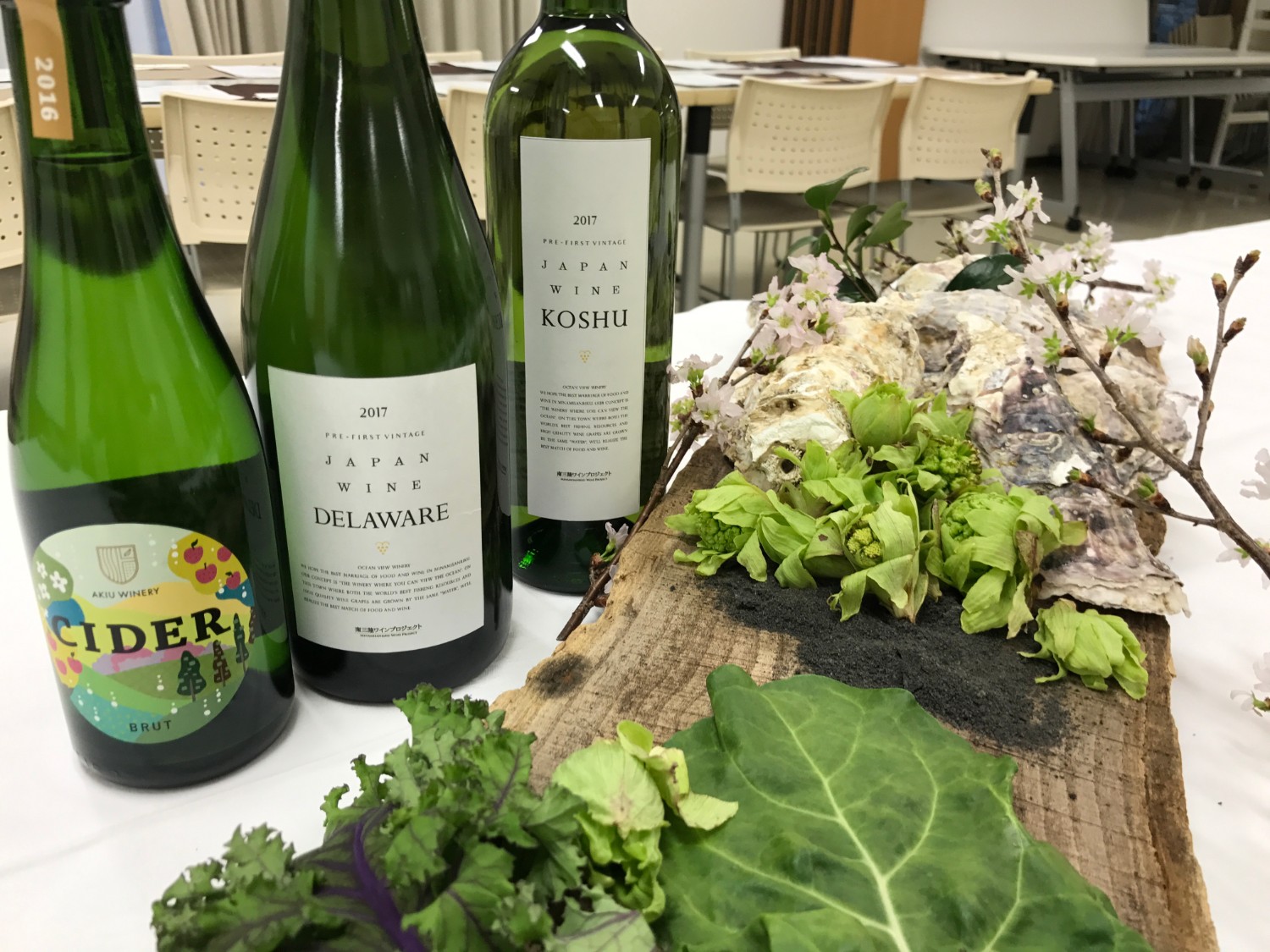 室内のテーブルの上に、2016年と2017年の日本産シードルとワイン3本が並べられ、その隣には木の板に乗せられた牡蠣の殻、ふきのとう、葉野菜、桜の枝が美しく盛り付けられている写真