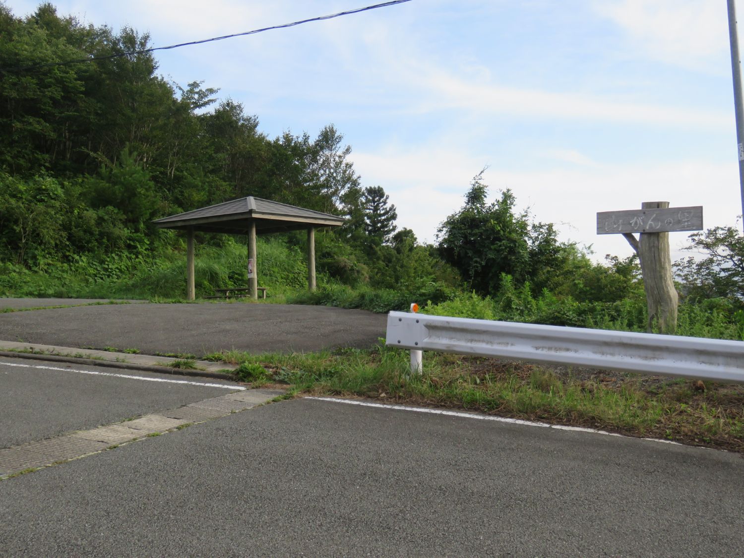 道路脇の広場に木の柱と屋根でできた四角い屋根の東屋があり、右側には「山がんの里」と書かれた木の看板が立っている写真