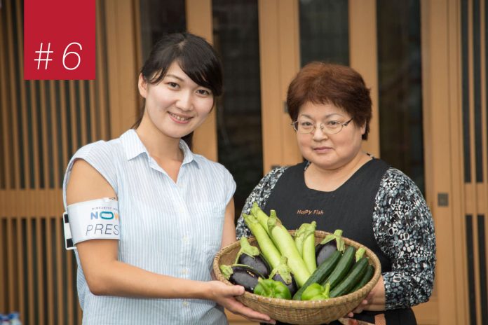 女性と黒色のエプロンを着用した女性が並んで立ち、収穫されたばかりのナスやキュウリ、ピーマン、白ナスなど色とりどりの夏野菜が入った丸いざるを持っている写真