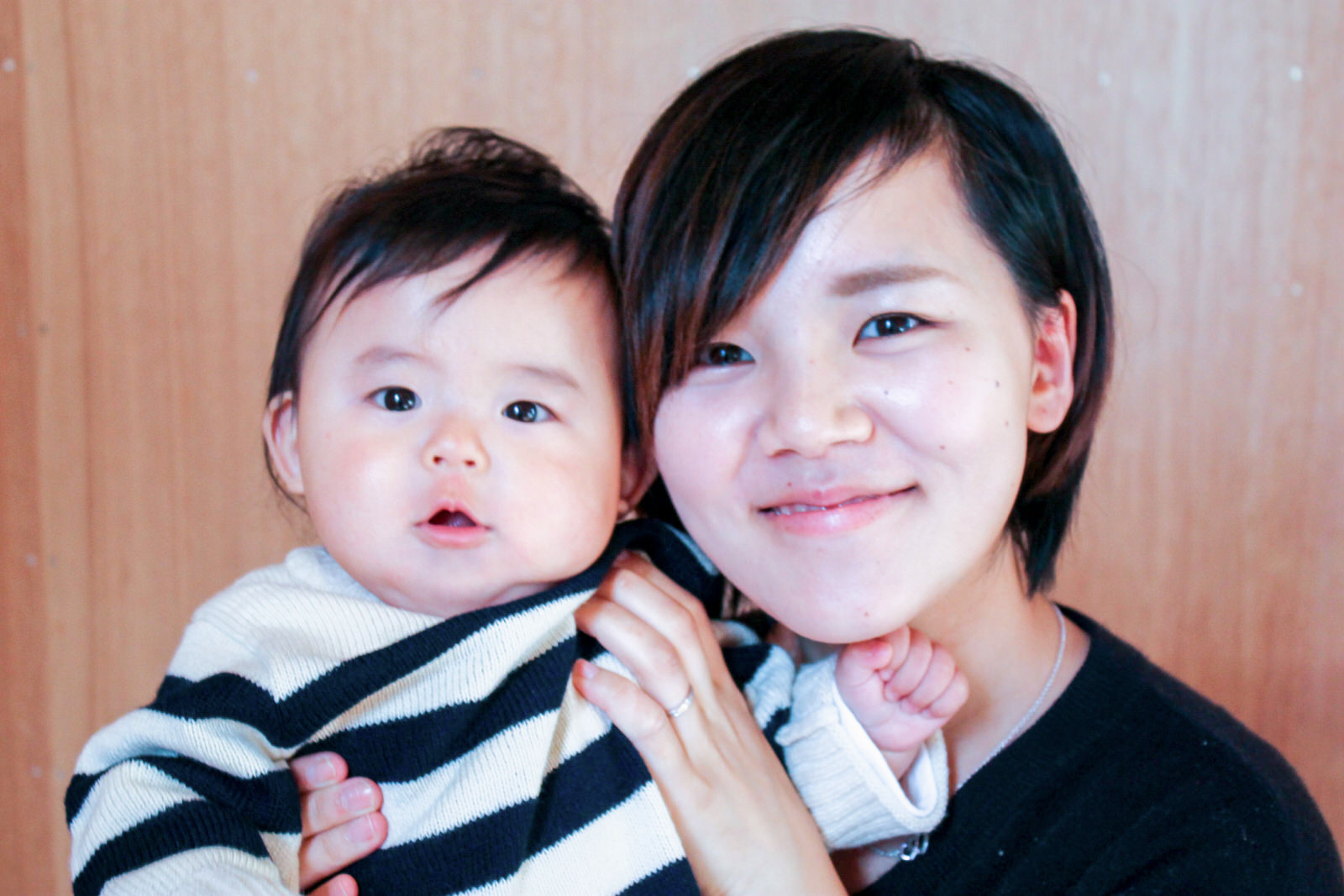 木の壁がある室内で、赤ちゃんを笑顔で抱きかかえている若い女性がカメラに向かって微笑んでいる写真