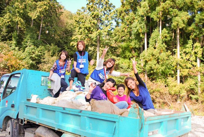 背の高い緑の木々が広がる明るい山間で、青い荷台のトラックの上に手を振ったりピースサインをしている5人の女性が乗っている写真