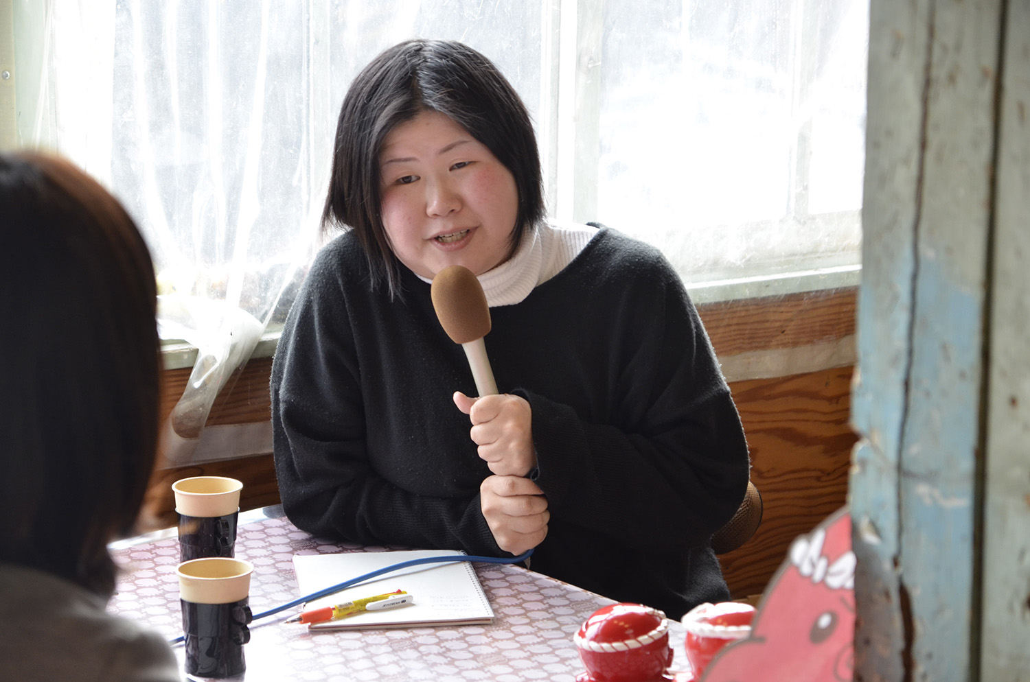 オクトパス君の置物が並んだテーブルの席に座った山内さんが、マイクを持ってインタビューを受けている様子の写真