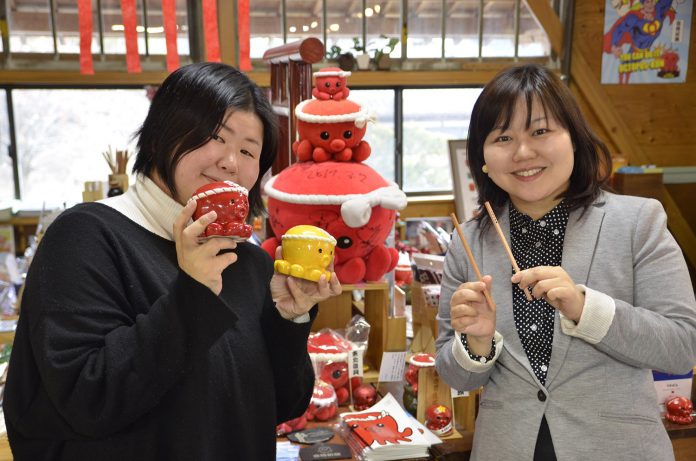 タコをモチーフにした「オクトパス君」のグッズが陳列された店内で、グッズを手に持った山内さんと女性リポーターが並んで立っている写真