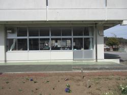 白い外壁と大きな窓が特徴の建物の一階部分で、室内には机や椅子が並び、外には花壇がある放課後児童クラブの写真