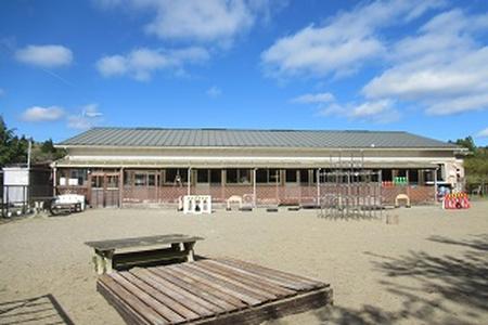 晴れた青空の下、広い砂地の園庭に木製遊具や鉄棒などが並ぶ、平屋建ての入谷ひがし幼児園の園舎を正面から写した写真