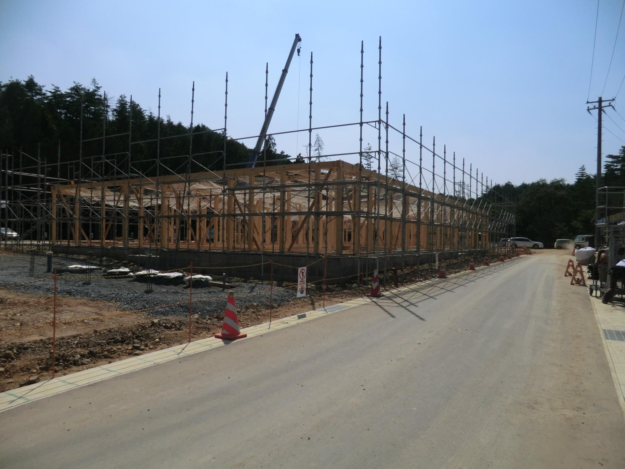 建設中の建物に足場が組まれ、木造の骨組みが整然と立ち上がる中、右側の舗装された道路沿いには工事車両や作業員が見られる写真