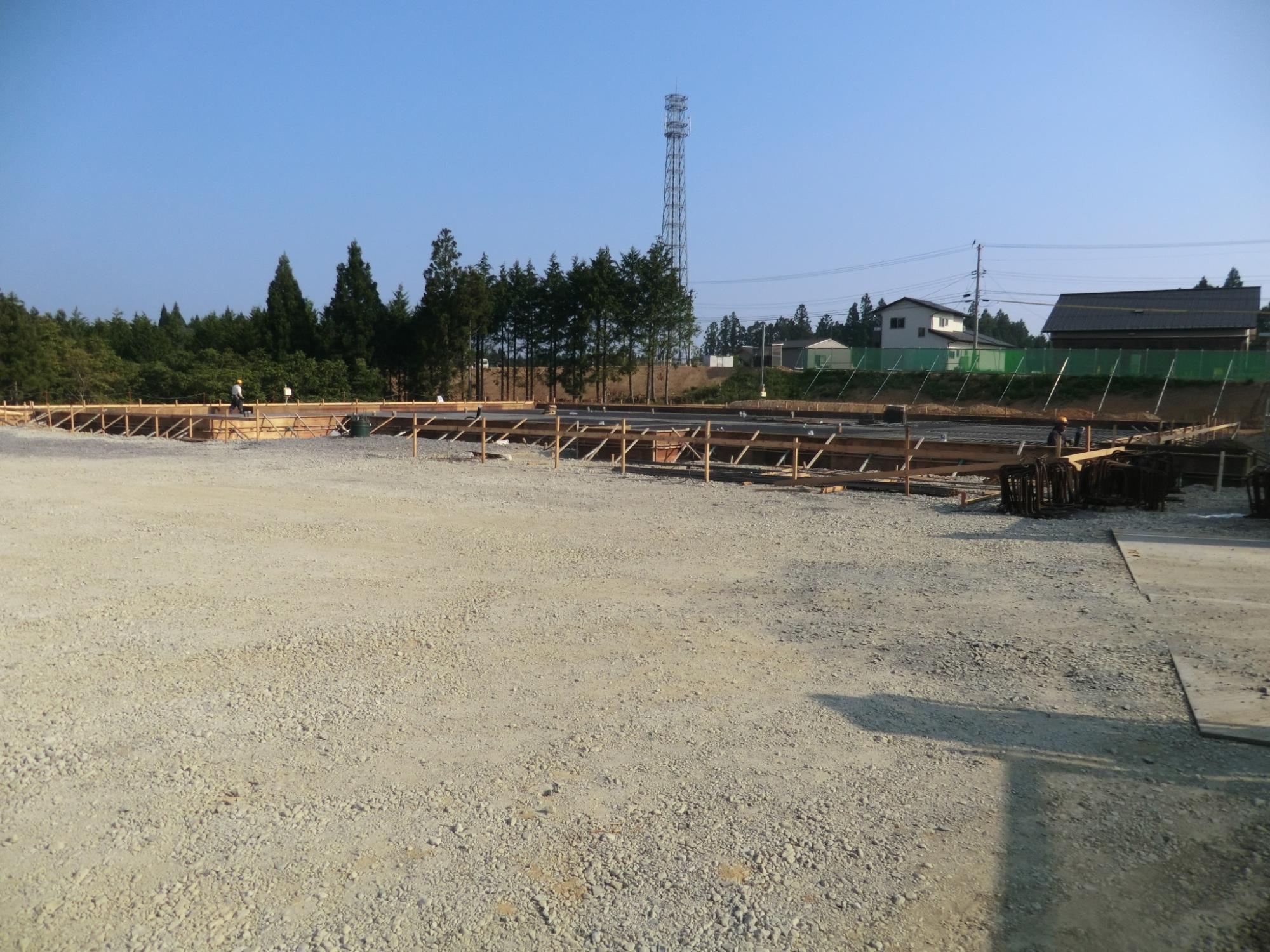 青空の下、広い砂利敷きの敷地で木枠が組まれ、数名の作業員が基礎工事を行っている建設現場の様子を捉えた写真