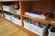 木の棚に白いプラスチック製のカゴが並び、積み木やままごとセットなどのおもちゃが整理されて収納されている様子を写した写真