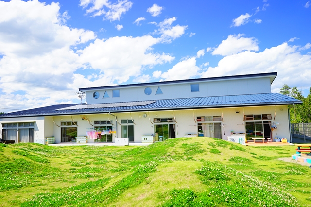 前面に起伏のある緑の芝生が広がり、開放感のある雰囲気の中、窓辺にはカラフルな装飾や遊具が置かれている白い外壁と青い屋根の伊里前保育所の外観写真