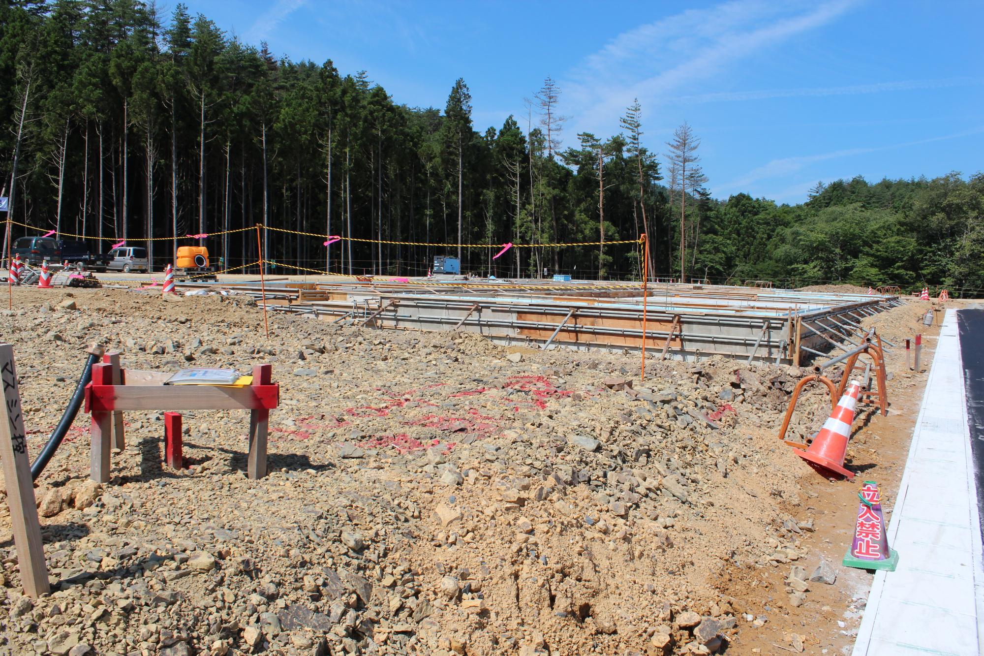 造成された広い土地に鉄枠が並び、複数の建物の基礎工事が同時に進められている様子が写され、奥には森林が広がり、手前には「立入禁止」の表示や工事用の資材が置かれている晴天の日の工事現場の写真