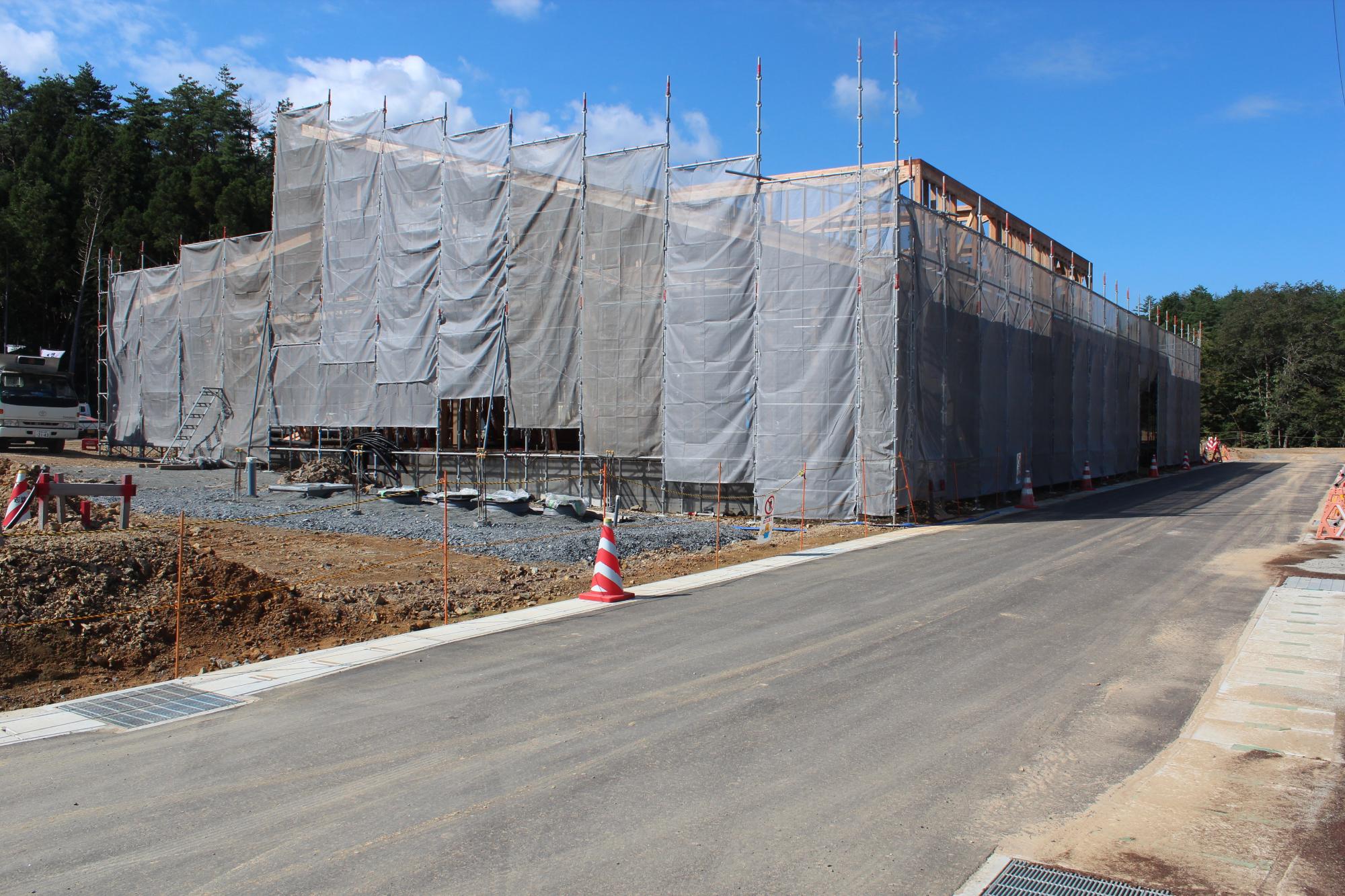 建設中の建物が足場と防塵用の白いシートで全面覆われ、道路に面して長く続くその構造物の内部では木造の梁や柱が組まれつつある様子が写された写真