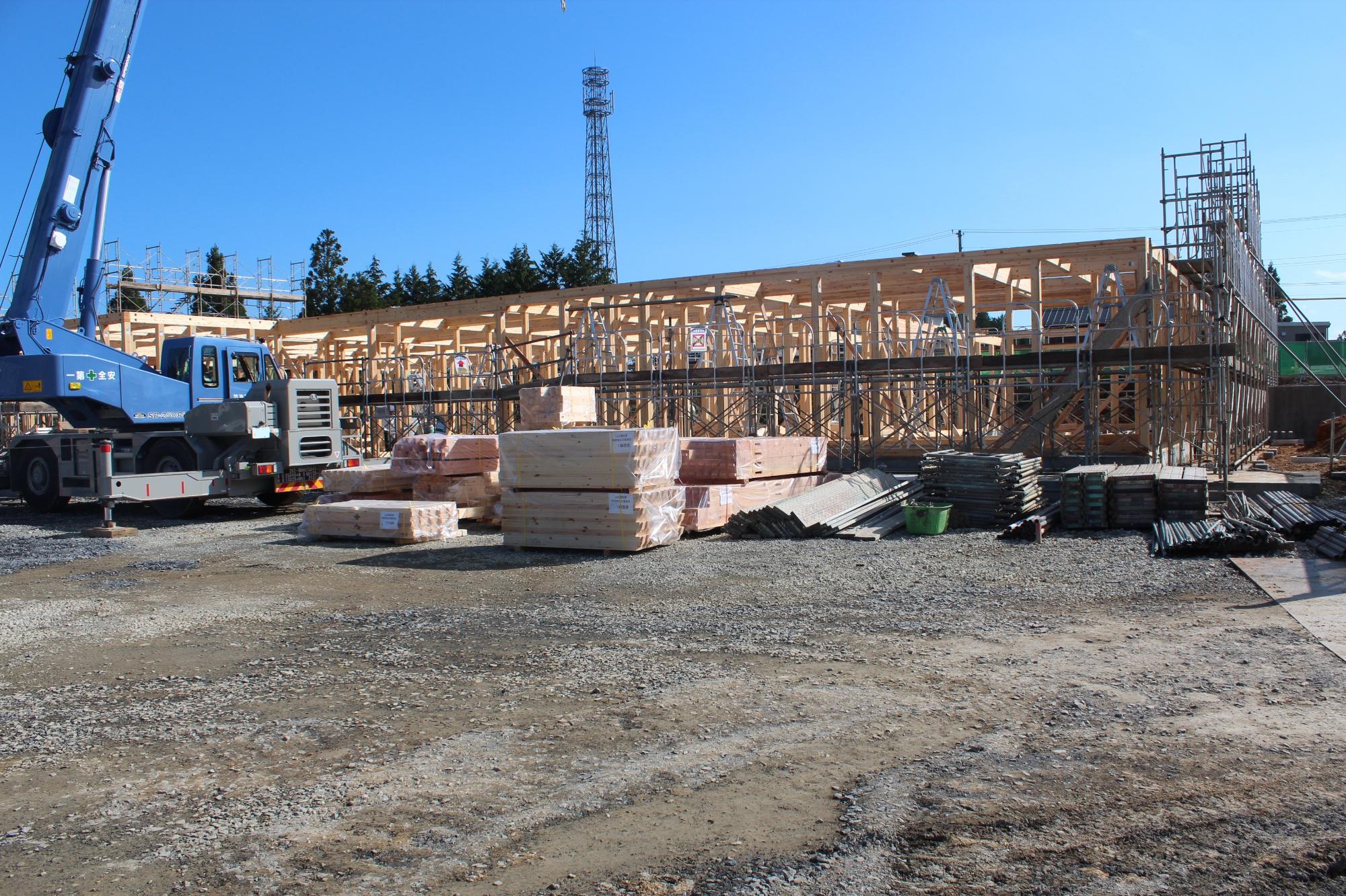 青空の下、木造建築の骨組みが組み上がりつつある建設現場で、クレーン車や足場、木材などの資材が整然と配置されている様子を写した写真