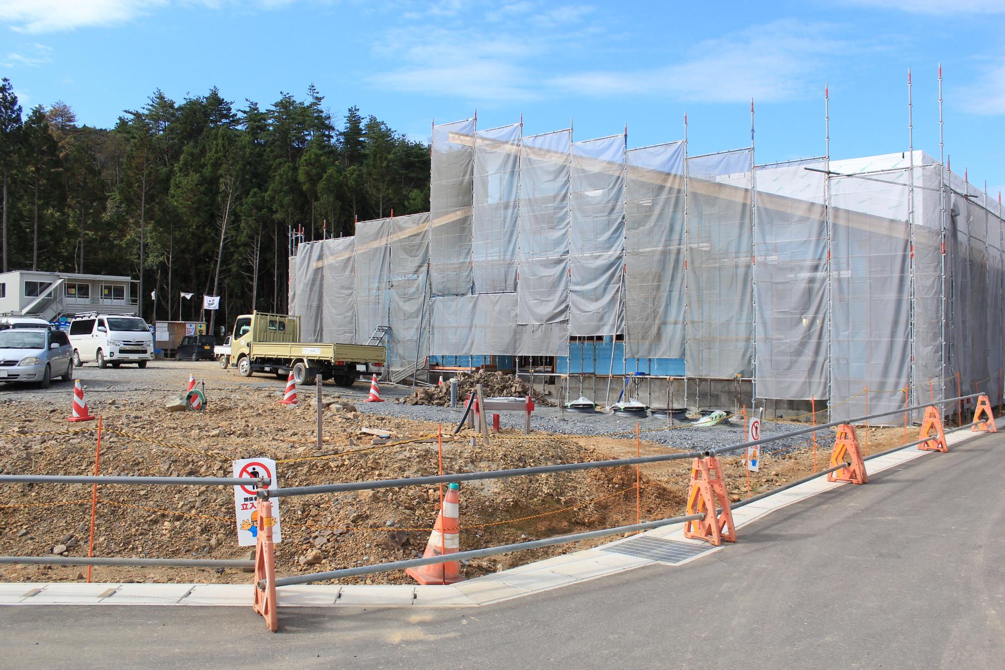 建設中の建物が足場と防塵用の白いシートで全面覆われ、晴天のもと整備された舗装道路や工事用の標識・カラーコーンが並ぶ様子が写された写真