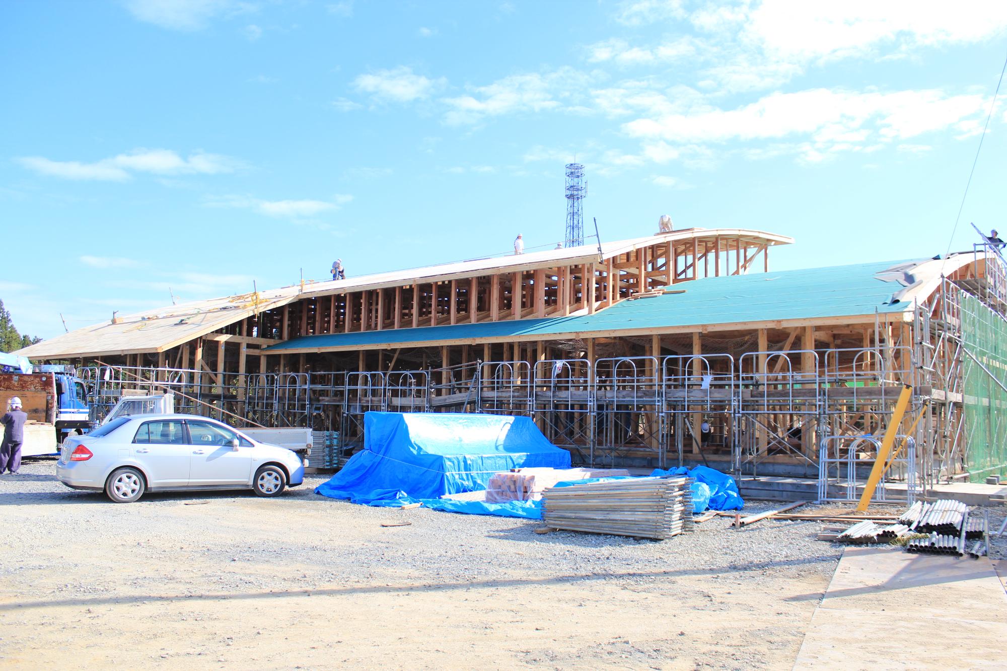 青空の下、屋根の骨組みが完成しつつある木造建築の現場で、足場やブルーシートに覆われた資材が並び、作業員が屋根上で作業を進めている様子を写した写真