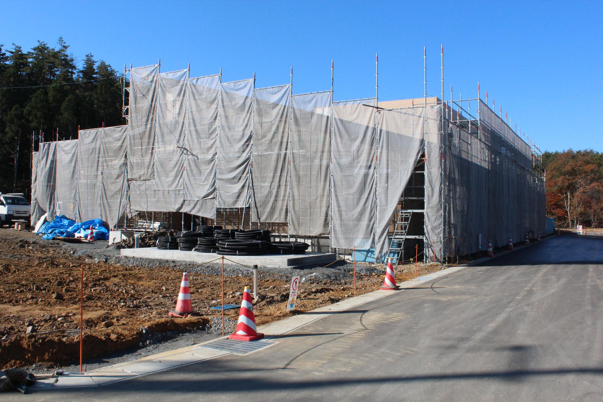 建設中の建物が足場と防塵用の白いシートで全面覆われ、建物の前に太くて黒い敗戦のような物が巻かれて置かれている写真