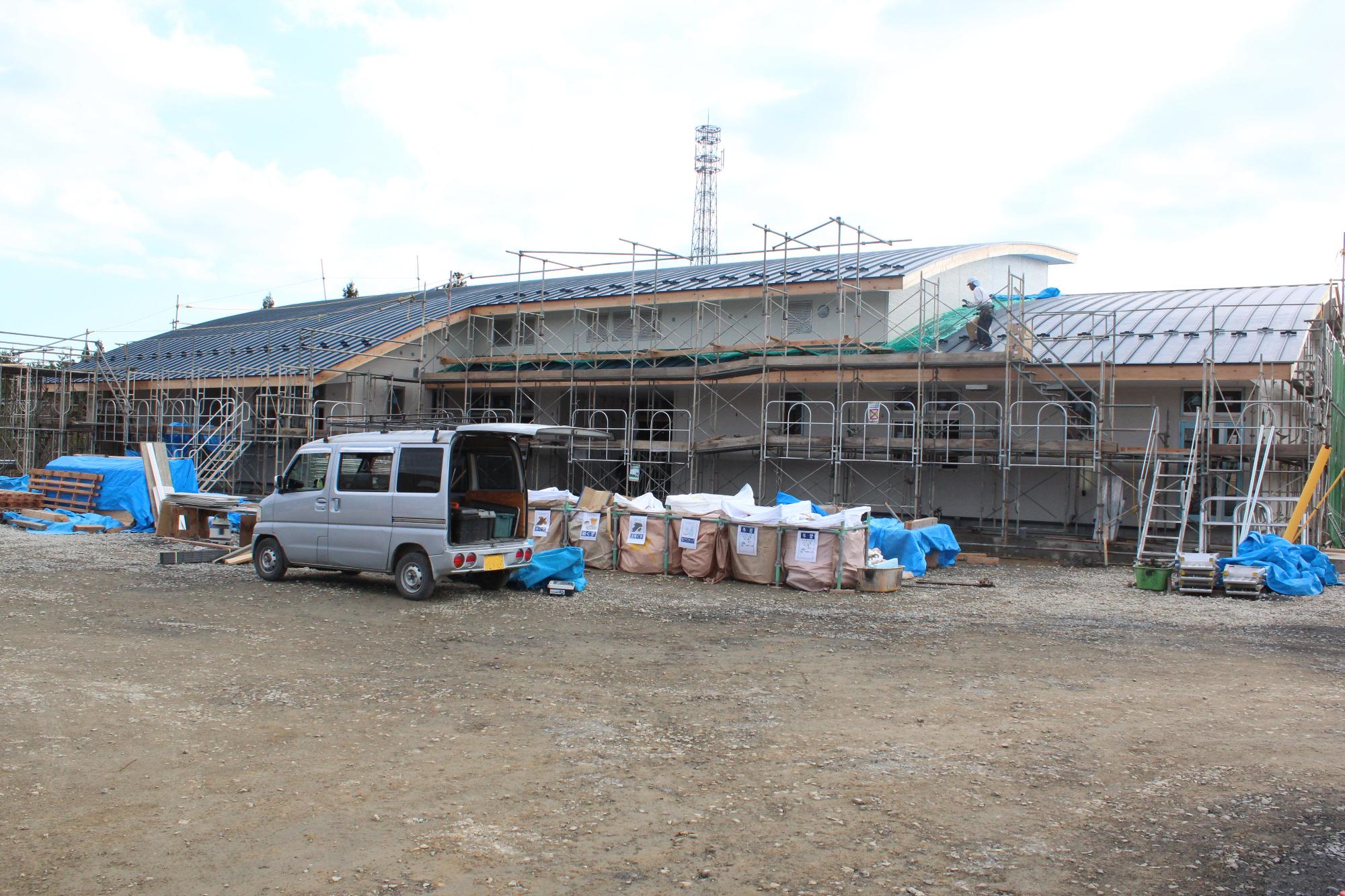 建物の外壁と屋根がほぼ完成し、足場が組まれた状態で作業員が屋根の上で作業している建設現場に、資材や工具を積んだ軽バンがとまっている様子を写した写真