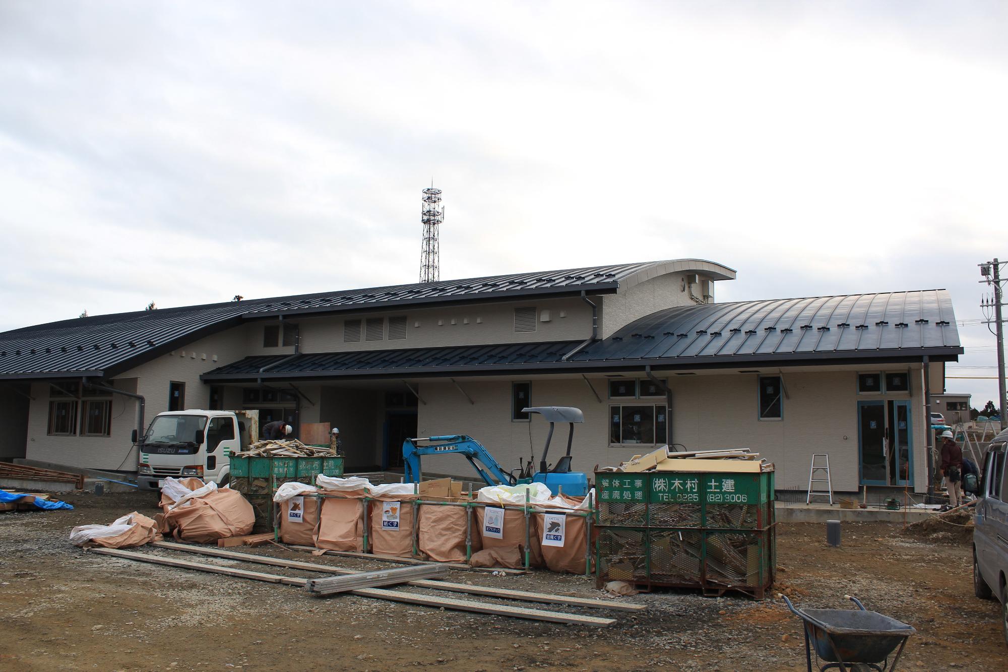 建物の外装と屋根がほぼ完成し、仕上げ工事が進められている現場で、前方には資材やごみ袋、トラックや小型重機が並び、数人の作業員が作業を行っている様子を写した写真