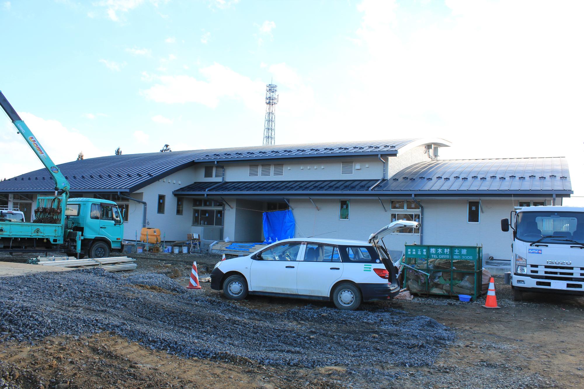 建設中の建物がほぼ完成しており、白い壁と黒い湾曲した屋根が特徴的な平屋の施設の前には工事用の車両や資材が並び、地面はまだ舗装されていない様子が見える写真