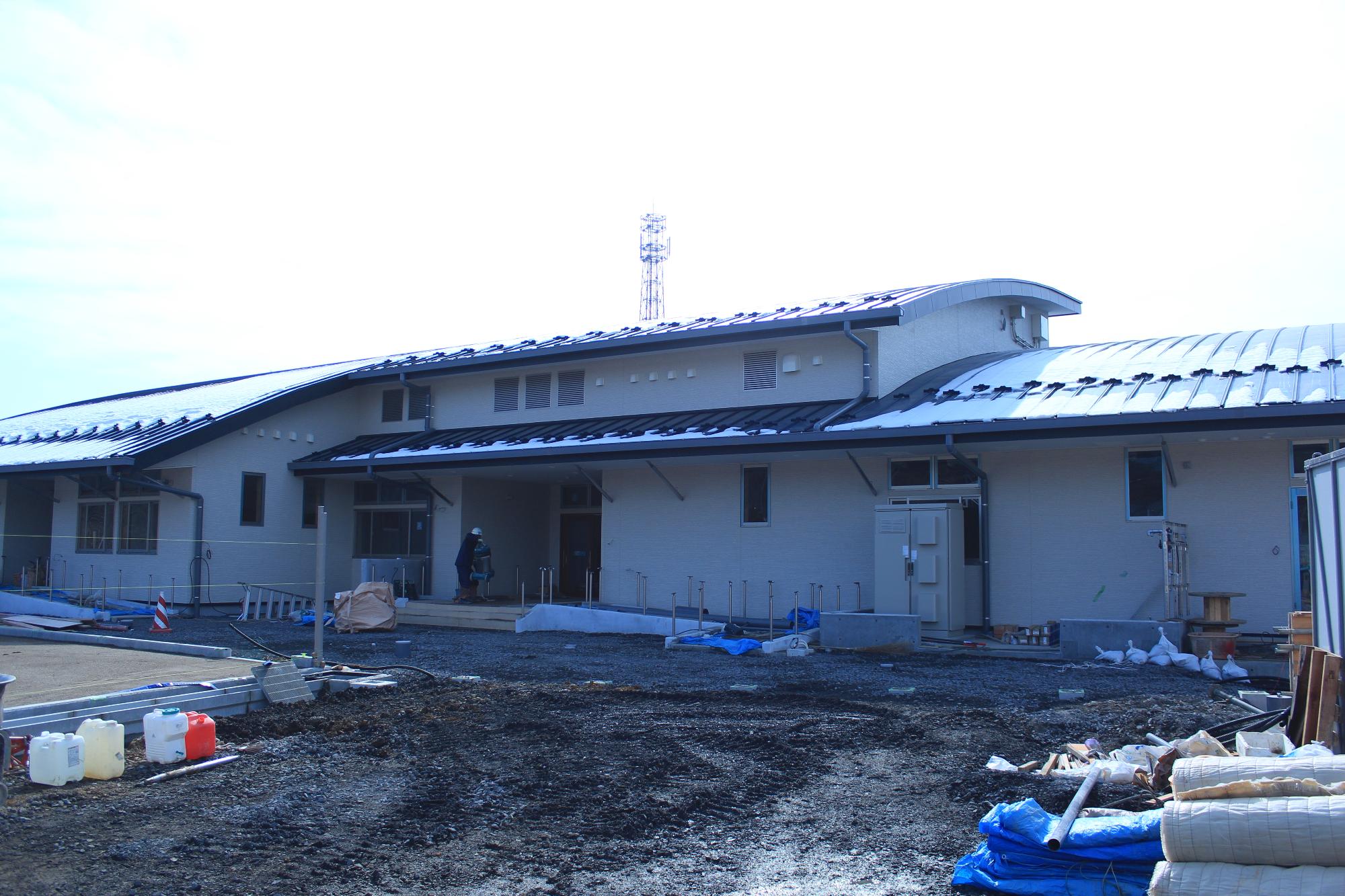 建設中の平屋の建物が完成に近づいており、白い外壁と湾曲した黒い屋根の一部に雪が積もり、正面入口には作業員が立ち、前の地面には建築資材や工具が置かれている写真