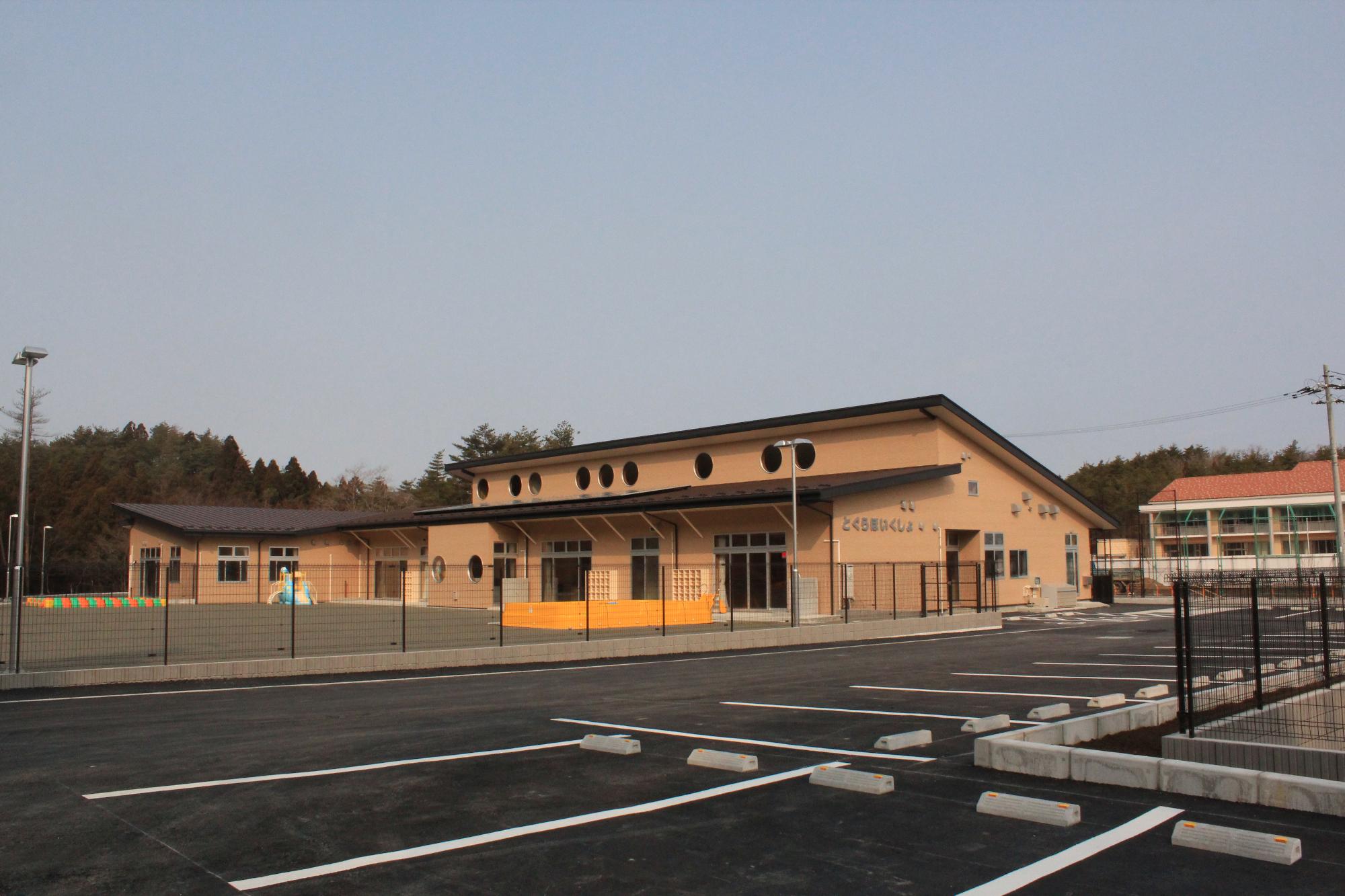 手前に駐車場が広がり、その奥にフェンスで囲まれた平屋建てで丸窓が取り付けられている戸倉地区子育て支援拠点施設の外観写真