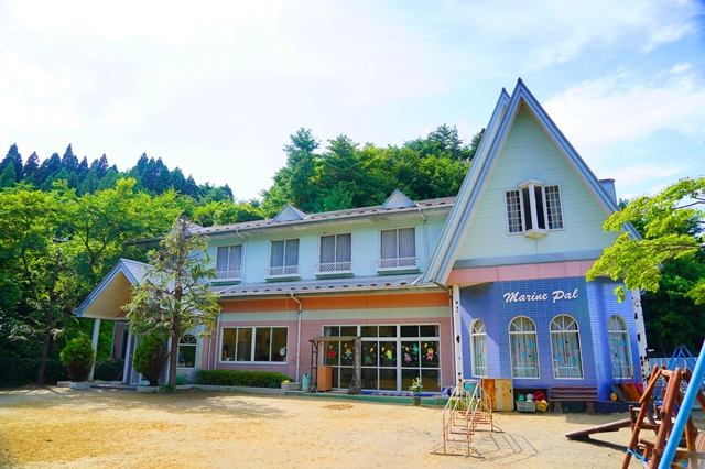 木々に囲まれた自然の中に建つ青とピンクの外壁を持つ二階建ての建物があり、右側には「Marine Pal」と書かれた看板が掲げられ、建物の前にはすべり台などの遊具が設置されているマリンパル保育園の外観写真