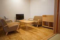 落ち着いたベージュのソファと一人掛けの椅子が木製のテーブルを囲むように配置され、テレビも置かれたシンプルな待合室の写真