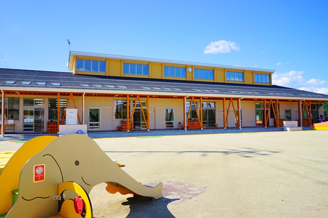幼稚園の園庭に面した明るい黄色の外壁の園舎と、手前の園庭に象の形をした遊具がある写真