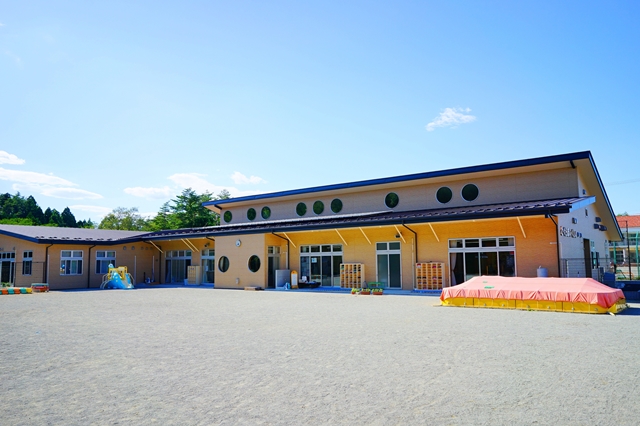 広い園庭と赤い砂場カバーが特徴的な、丸窓のある明るいデザインの保育園の外観写真
