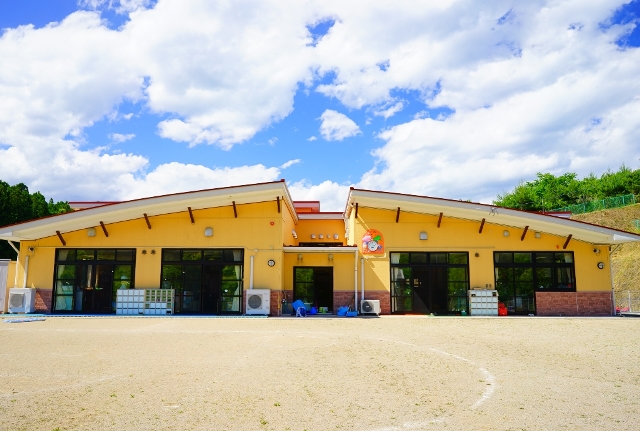青空の下、広い園庭に面した黄色い建物が左右対称に建っており、窓が大きく開放感がある名足こども園の外観写真
