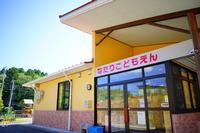 黄色い外壁の建物の玄関上に「なたりこどもえん」と書かれた赤い文字の看板が掲げられ、保育施設の正面入口が青空の下で明るく映し出されている写真