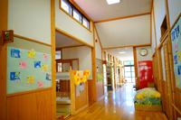 木のぬくもりを感じる明るい保育園の廊下に、子どもたちの手形付きの鯉のぼりの形をした色紙が壁に飾られ、奥にはクラスルームやロッカーが並んでいる写真