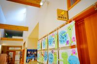 木製の天井と壁に囲まれた明るい廊下の一角に、かわいらしい青いカエルのキャラクターが描かれた子どもたちの作品が壁に並べられて展示されている写真