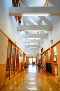 白い梁と木の床が美しい、奥まで長く続く廊下に教室の入口が並ぶ保育園内の写真