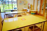 明るい日差しが差し込むガラス窓のそばに、黄色い天板の机と小さな椅子が並べられ、奥にはおままごと用のキッチンや棚が整然と配置されている保育園の室内を写した写真